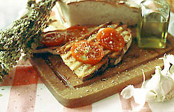 Bruschetta mit Tomaten auf einem Holzbrett, umgeben von Thymian, einem angeschnittenen Laib Brot, Olivenöl und Knoblauch